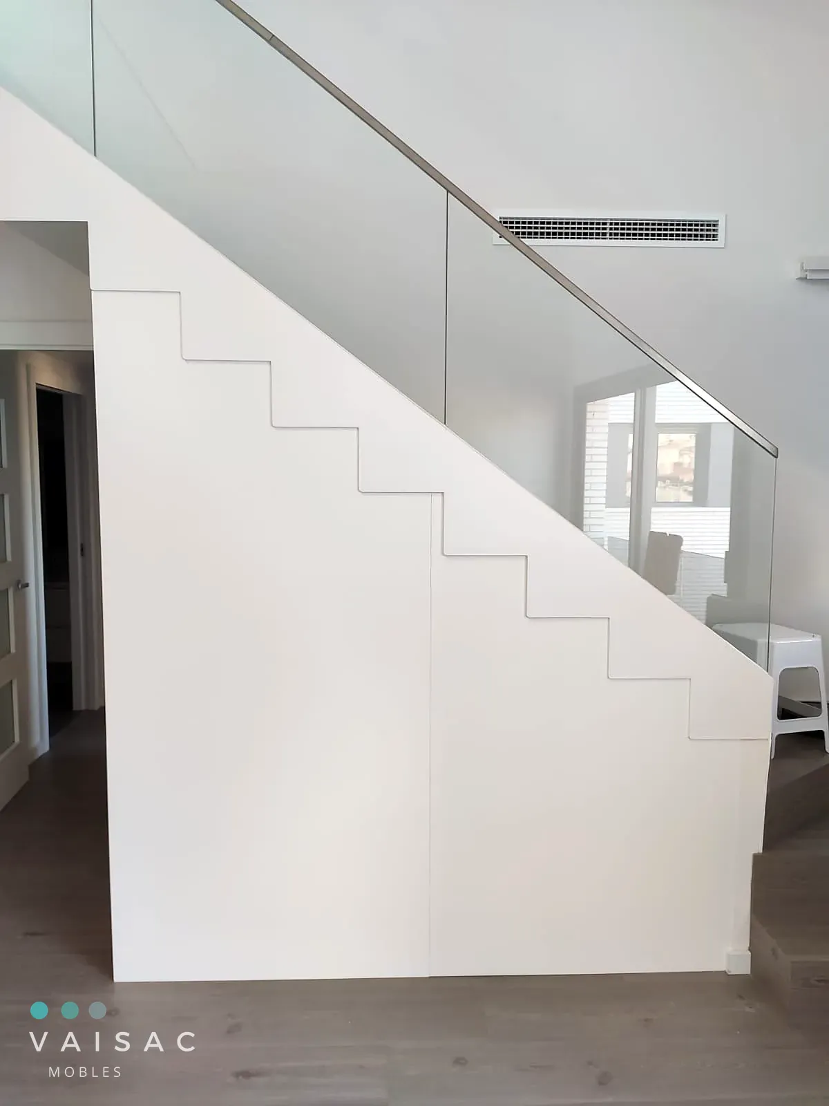 Armario empotrado bajo escalera con frentes blancos lisos sin tiradores en una escalera moderna con barandilla de cristal